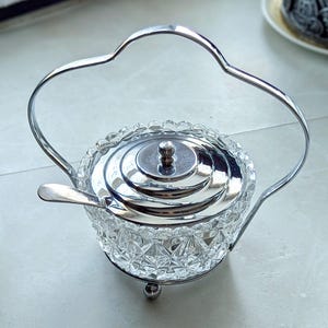 May include: A silver-plated sugar bowl with a clear, faceted glass base and a matching lid. The bowl has a small spoon and a handle. The sugar bowl is a decorative item.