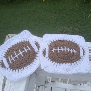 Könnte beinhalten: Zwei handgefertigte Häkeluntersetzer. Jeder Untersetzer hat ein braunes Football-Design mit weißen Nähten, das auf einem weißen quadratischen Rand angeordnet ist. Die Untersetzer sind auf einer weißen Holzoberfläche mit grünem Hintergrund platziert.
