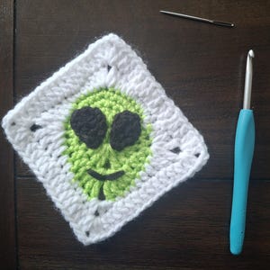 May include: A handmade crochet square featuring an alien face design. The alien has black eyes and a smiling mouth, set against a green background and a white border. A blue crochet hook and a needle are also in the image.
