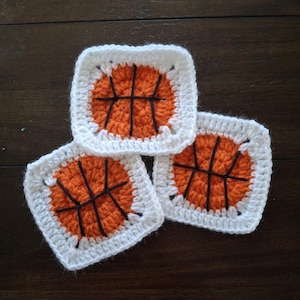 Puede incluir: Tres cuadrados tejidos a crochet con un diseño de baloncesto. Los cuadrados son blancos con una pelota de baloncesto naranja en el centro y costuras negras para las líneas de la pelota de baloncesto.