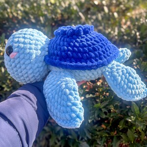 May include: A handmade crochet turtle in shades of blue. The turtle has a light blue head and limbs, a darker blue shell, and black eyes with a pink mouth. The plush toy is soft and textured, perfect for a child's room or as a decorative item.