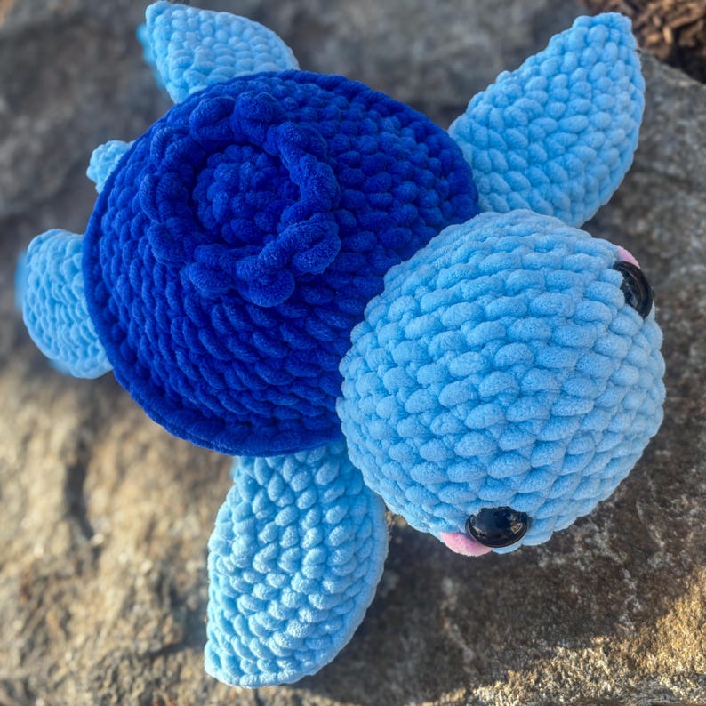May include: A handmade plush turtle in shades of blue. The turtle has a dark blue shell, light blue flippers, and a light blue head. The eyes are black, and the mouth is pink. The turtle is made of a soft, textured material.