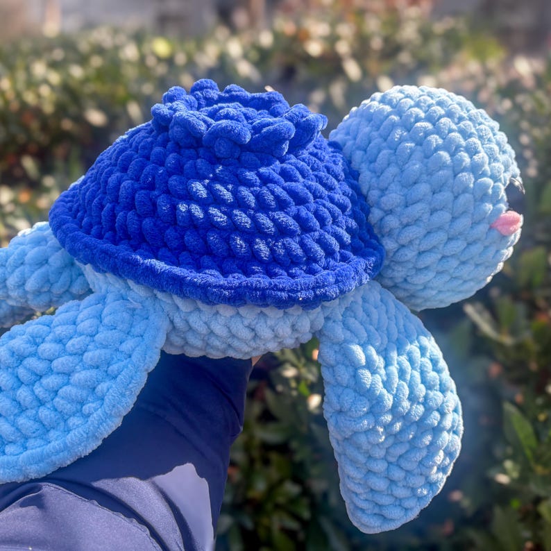May include: A handmade plush turtle in shades of blue. The turtle has a dark blue shell, light blue flippers, and a light blue head. The turtle's face has small black eyes and a pink mouth. The plush toy is made of a soft, textured material.