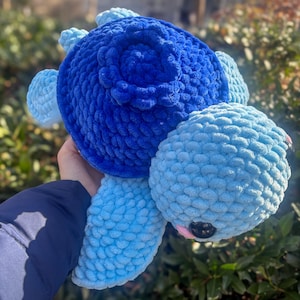 May include: A handmade plush turtle in shades of blue. The turtle features a large, textured shell in a deep blue color, with lighter blue flippers and head. The eyes are black, and the mouth is a soft pink. The plush toy is made of a soft, chunky yarn.