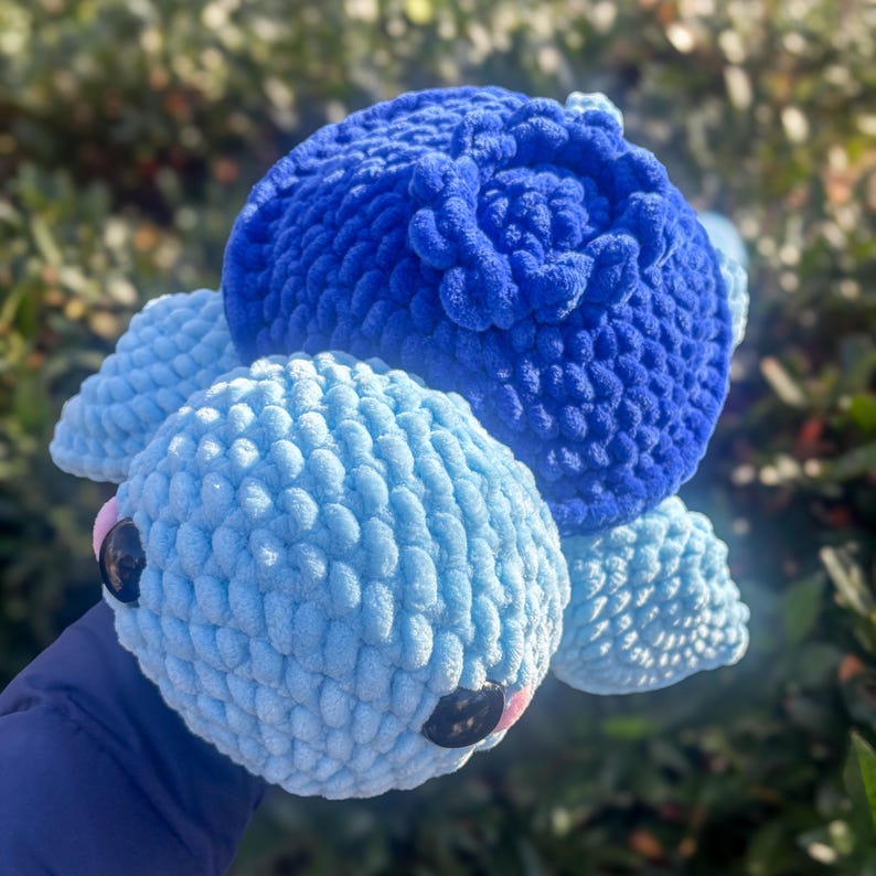 May include: A handmade, plush turtle toy in shades of blue. The turtle features a large, textured head in deep blue, a lighter blue body, and four flipper-like appendages. The eyes are black, and the mouth is pink.