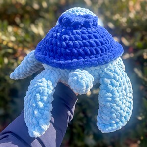 May include: A handmade, plush turtle in shades of blue. The turtle features a dark blue shell, light blue flippers, and a light blue head. The soft, textured material gives the turtle a cuddly appearance. The turtle is a handcrafted item.