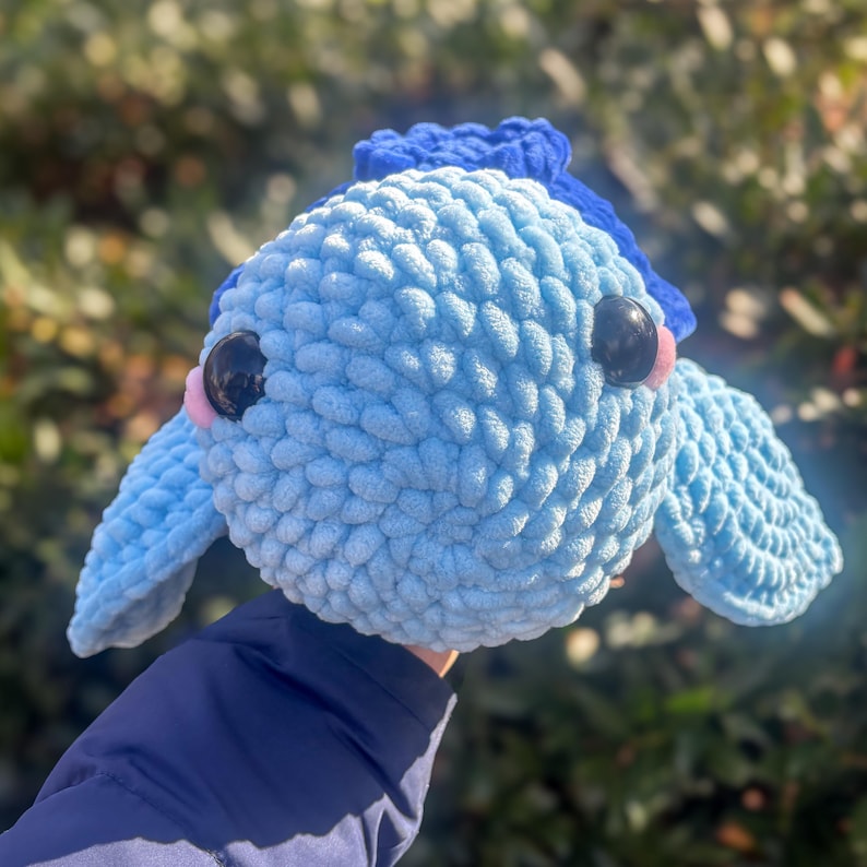 May include: A handmade, light blue crochet fish with dark eyes and pink accents. The plush toy has a textured, fluffy appearance and features two small fins and a dark blue fin on top. The fish is a whimsical, soft toy.