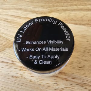 May include: A black container of UV Laser Framing Powder. The lid has white text that reads: Enhances Visibility, Works On All Materials, and Easy To Apply & Clean. The container sits on a light brown wooden surface.