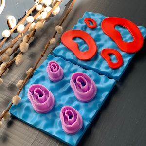 Puede incluir: Un juego de cortadores de galletas de color rosa y rojo en forma de óvalo ondulado. Los cortadores están sobre una estera de horneado de silicona azul.