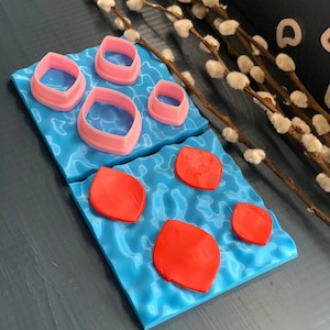 Op de afbeelding: Een set roze en rode siliconen fondant-uitsteekvormen en een blauwe siliconen mat met een waterrimpelontwerp. De uitsteekvormen zijn rond rechthoekig gevormd en de mat heeft drie rode fondantvormen erop.