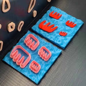 Puede incluir: Dos alfombras azules con cortadores de galletas de plástico rosa y rojo en forma de peine. Los cortadores están dispuestos en diferentes tamaños en las alfombras.