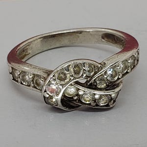 Puede incluir: Un anillo plateado con diseño de nudo, incrustado con piedras preciosas transparentes. El anillo presenta una banda con pequeñas piedras y un grupo más grande de piedras en el centro del nudo. Se encuentra sobre una superficie gris claro.