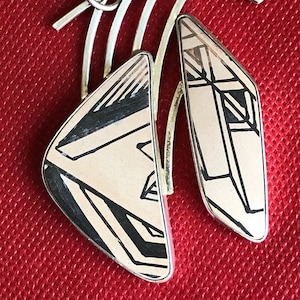 Könnte beinhalten: Ein silberner Anhänger mit zwei weißen und schwarzen geometrischen Mustern. Der Anhänger ist an einer silbernen Öse mit einer Schlaufe befestigt.