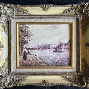 May include: A framed painting depicting a scene of a bridge over a river with buildings in the background. Two people and a dog are walking along the riverbank.