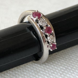 Peut inclure: Une bague en argent avec une rangée de cinq pierres précieuses rouges et de petites pierres blanches serties entre elles. La bague est posée sur une surface noire.