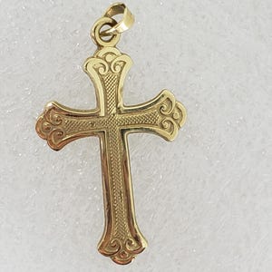 May include: A gold-colored pendant in the shape of a cross. The cross features ornate detailing and a loop at the top for a chain. The design includes scrolled edges and textured patterns, creating a classic and elegant look. The pendant is likely made of gold.