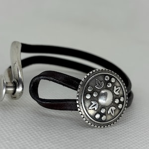 May include: A black leather bracelet with a decorative silver button clasp. The button features a detailed design with leaf and dot patterns. The bracelet is designed with two leather bands and a silver hook.