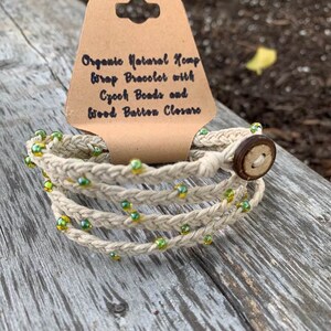 May include: A natural hemp wrap bracelet with green Czech beads and a wood button closure. "Organic Natural Hemp Wrap Bracelet with Czech Beads and Wood Button Closure" is written on a brown tag.