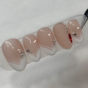 May include: Five oval-shaped fake nails with a light pink base and white tips. The nails have a white V-shaped design at the base and are decorated with small rhinestones. One nail has a gold chain design and a red rhinestone.