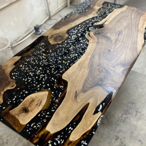 Könnte beinhalten: Rechteckiger Holztisch mit glänzendem Finish. Der Tisch hat auf einer Seite eine natürliche Holzmaserung und auf der anderen Seite schwarzes Epoxidharz mit Gold- und Silberflocken. Ein einzigartiges Möbelstück, ca. 150 cm lang.