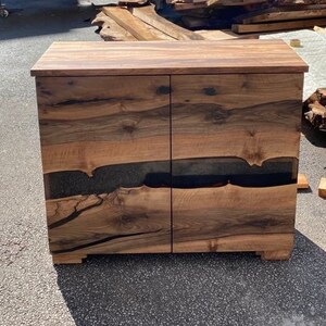 May include: A wooden cabinet with two sets of doors. The cabinet is made of dark wood with a black resin inlay. The cabinet is sitting outside in front of a wooden building. The building has the text "Lara Wood's" on it.