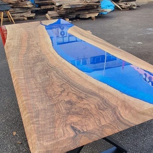May include: A handcrafted wooden table with a blue epoxy resin river. The table has a natural wood grain finish and is supported by a black metal base. The table measures approximately 72 inches long.