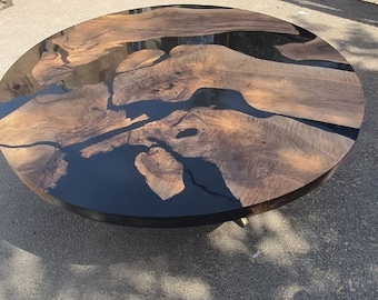 May include: A round wooden coffee table with a dark epoxy resin inlay. The table features a natural wood grain pattern with black resin filling the gaps, creating a unique design. The table is set on a dark metal base.