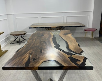 May include: A large, rectangular wooden table with a dark brown wood grain and a clear epoxy river. The table has a silver metal X-shaped base. Another table is visible in the background.