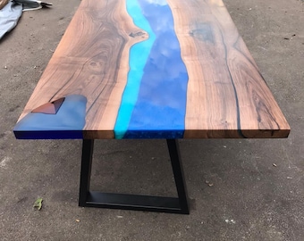 Puede incluir: Una mesa rectangular de madera con un río de resina azul en el centro. La mesa tiene una veta de madera oscura y se apoya sobre patas de metal negro. El río de resina es un degradado de azules.
