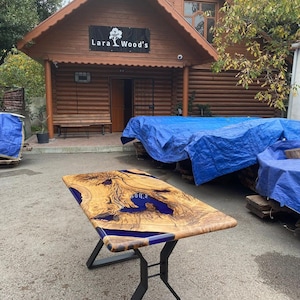 Könnte beinhalten: Ein rechteckiger Holztisch mit natürlicher Kante und blauen Harzakzenten. Der Tisch hat ein natürliches Holzmaserungsmuster und steht auf schwarzen Metallbeinen. Vor einem Holzgebäude mit dem Text "Lara Wood's".