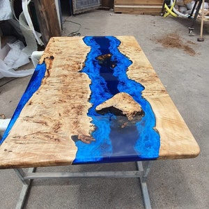 Op de afbeelding: Een rechthoekige houten tafel met een rivierontwerp. De tafel heeft een lichtgekleurd houten blad met helderblauwe epoxyhars die door het midden stroomt. Het wordt ondersteund door een zilverkleurig metalen frame. De tafel is ongeveer 150 cm lang.