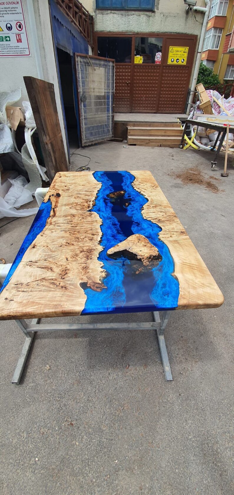 Op de afbeelding: Een rechthoekige houten tafel met een rivierontwerp. De tafel heeft een lichtgekleurd houten blad met helderblauwe epoxyhars die door het midden stroomt. Het wordt ondersteund door een zilverkleurig metalen frame. De tafel is ongeveer 150 cm lang.