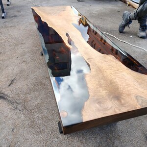 May include: A rectangular wooden table with a clear epoxy resin finish. The wood has a natural grain pattern with a light brown colour. The resin creates a reflective surface, and the table is supported by dark legs.