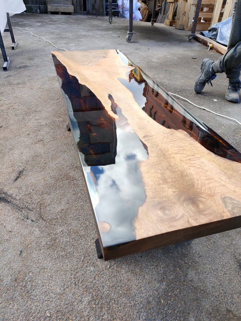 May include: A rectangular wooden table with a clear epoxy resin finish. The wood has a natural grain pattern with a light brown colour. The resin creates a reflective surface, and the table is supported by dark legs.