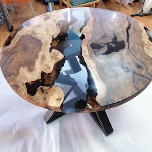 May include: A round wooden table with a unique design. The tabletop features a combination of light and dark wood tones with a clear, glossy resin inlay. The table has a black, geometric base. The table is approximately 48 inches in diameter.