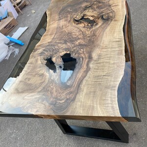 May include: A large, live-edge wooden table with a natural, light brown wood grain. The table has a clear epoxy resin finish and sits on a black metal base. The table's dimensions are approximately 96 inches long and 40 inches wide.
