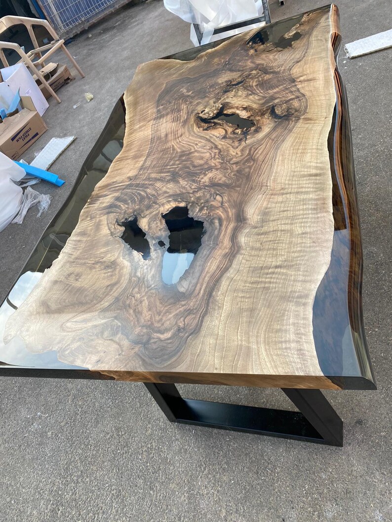 May include: A large, live-edge wooden table with a natural, light brown wood grain. The table has a clear epoxy resin finish and sits on a black metal base. The table's dimensions are approximately 96 inches long and 40 inches wide.