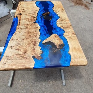 Op de afbeelding: Een rechthoekige houten tafel met een rivier van blauwe epoxyhars in het midden. Het hout heeft een lichtbruine kleur met donkerdere bruine patronen. De tafel wordt ondersteund door zilveren metalen poten.
