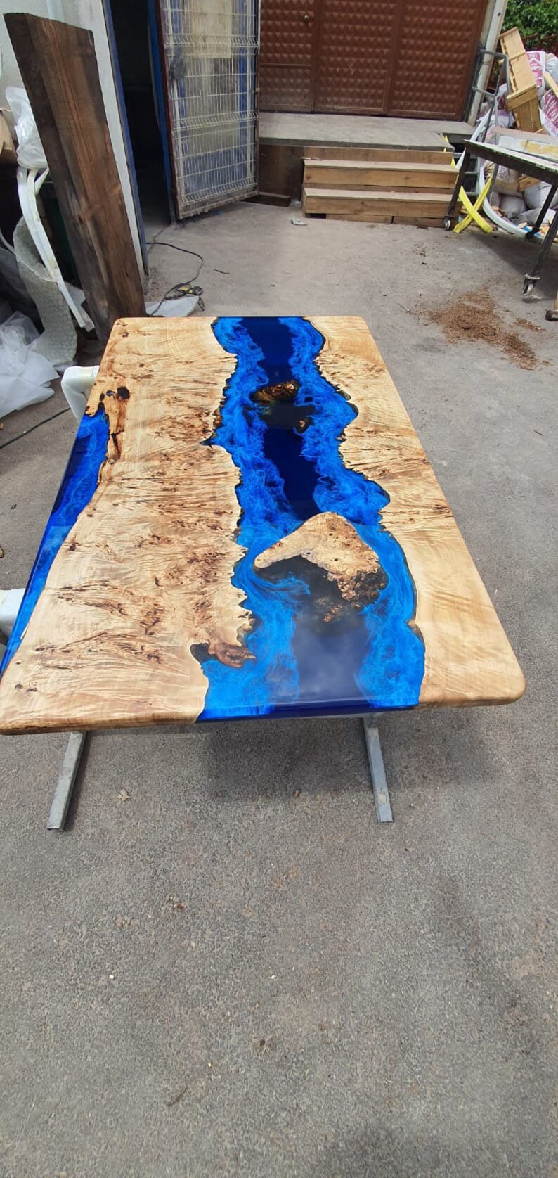 Op de afbeelding: Een rechthoekige houten tafel met een rivier van blauwe epoxyhars in het midden. Het hout heeft een lichtbruine kleur met donkerdere bruine patronen. De tafel wordt ondersteund door zilveren metalen poten.