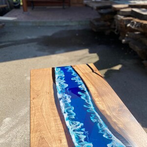 May include: A wooden coffee table with a river of blue resin. The resin is a vibrant blue with white accents, resembling ocean waves. The table's wooden surface has a natural grain and a warm, light brown color.