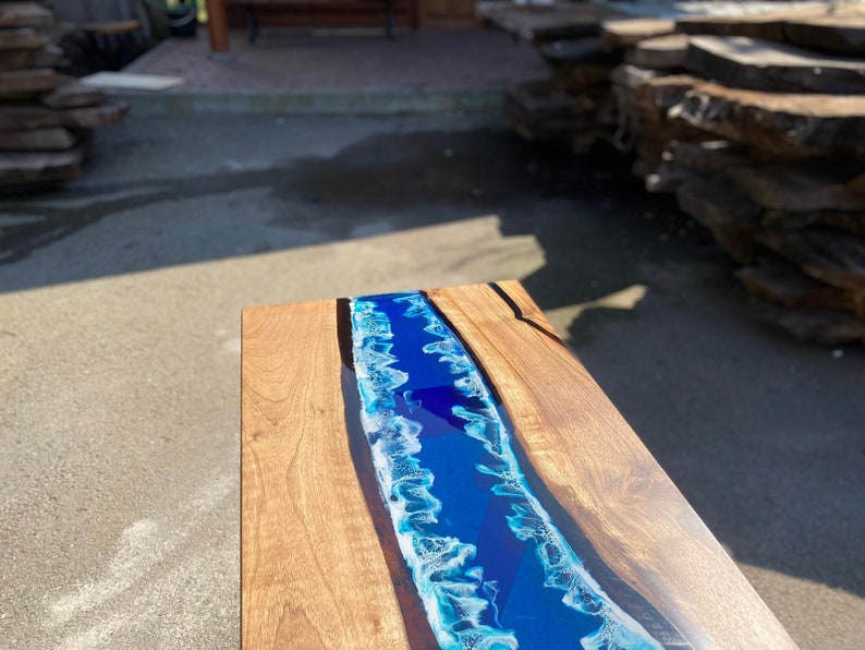 May include: A wooden coffee table with a river of blue resin. The resin is a vibrant blue with white accents, resembling ocean waves. The table's wooden surface has a natural grain and a warm, light brown color.