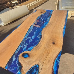 May include: A rectangular wooden table with a blue epoxy resin river design. The table features natural wood grain and a glossy finish. The blue resin has white swirls, creating a water-like effect. The table is approximately 72 inches long.