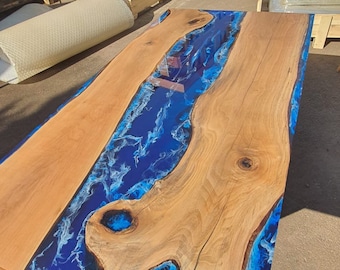May include: A rectangular wooden table with a blue epoxy resin river design. The table features natural wood grain and a glossy finish. The blue resin has white swirls, creating a water-like effect. The table is approximately 72 inches long.