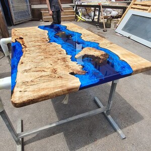 Op de afbeelding: Een grote, rechthoekige houten tafel met een rivierachtige blauwe epoxyharsinleg. De tafel wordt ondersteund door een zilverkleurig metalen frame. Het hout heeft een lichtbruine kleur met unieke nerfpatronen.