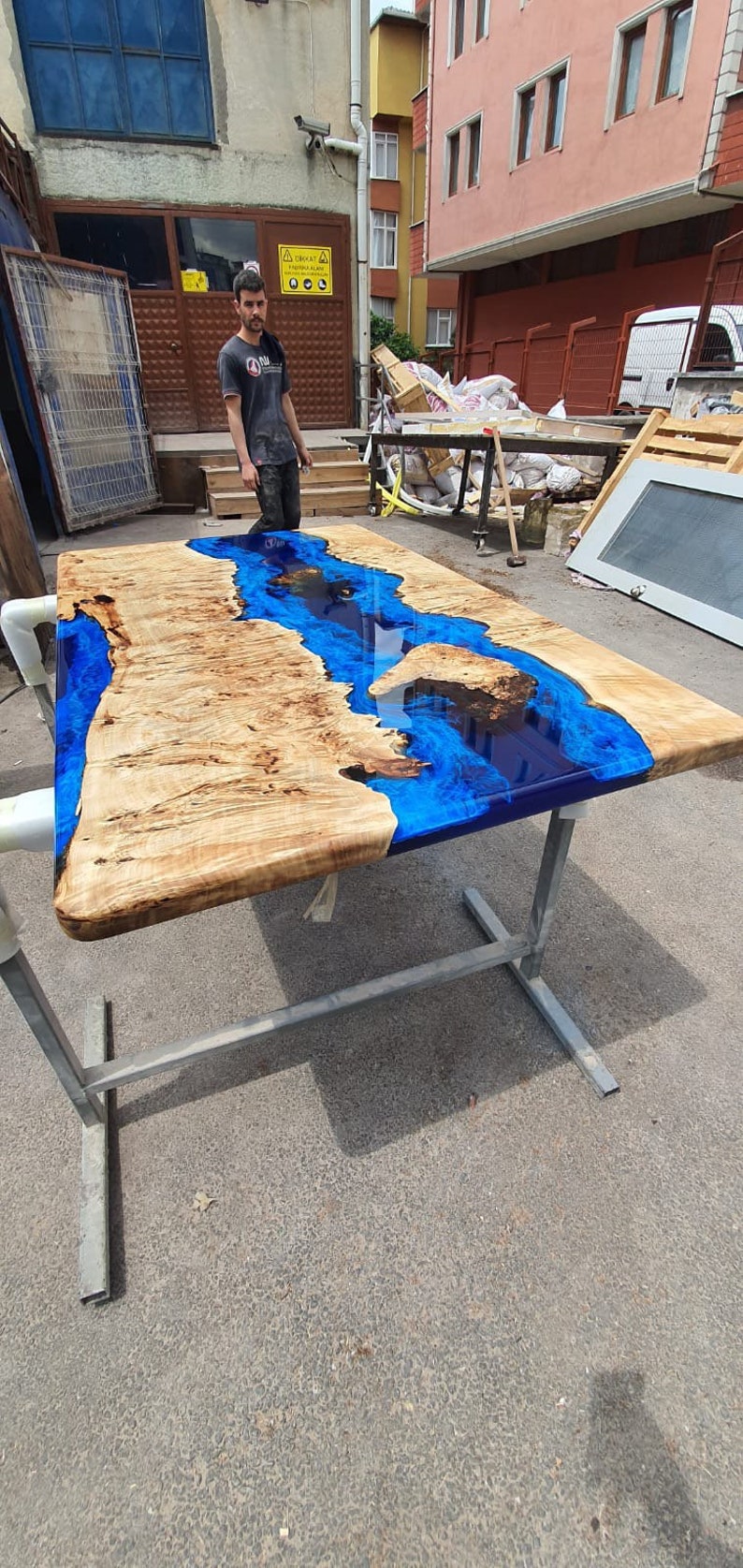 Op de afbeelding: Een grote, rechthoekige houten tafel met een rivierachtige blauwe epoxyharsinleg. De tafel wordt ondersteund door een zilverkleurig metalen frame. Het hout heeft een lichtbruine kleur met unieke nerfpatronen.