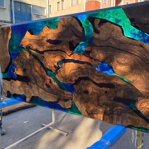 Op de afbeelding: Een grote, rechthoekige houten tafel met een uniek ontwerp. De tafel heeft een donkerbruine houtnerf met secties gevuld met levendig blauwe en groene epoxyhars. De tafel wordt ondersteund door een metalen frame.