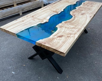 May include: A rectangular wooden table with a blue epoxy river design. The table has a light-colored wood top with a blue, river-like epoxy center. It is supported by black metal legs. The table is approximately 72 inches long.