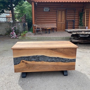 Puede incluir: Un cofre de madera con acabado de madera natural y un diseño de río de resina epoxi gris oscuro. El cofre se apoya sobre patas de metal negro. El letrero de la tienda dice "Lara Wood's".