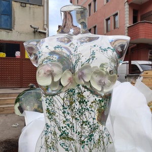 Puede incluir: Busto de resina transparente con flores secas y vegetaci&oacute;n incrustadas. El busto presenta una forma femenina con flores blancas y moradas similares a orqu&iacute;deas. La escultura se exhibe al aire libre.