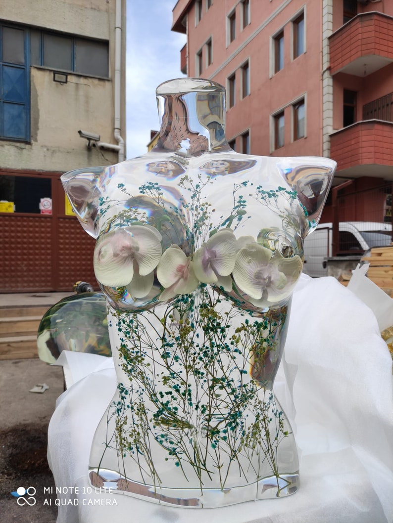 Puede incluir: Busto de resina transparente con flores secas y vegetaci&oacute;n incrustadas. El busto presenta una forma femenina con flores blancas y moradas similares a orqu&iacute;deas. La escultura se exhibe al aire libre.